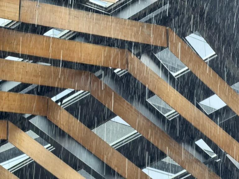 营地某日下午透过木格栅往外拍的暴雨，斜向密集雨柱，窗外是阴云下的现代高层公寓楼