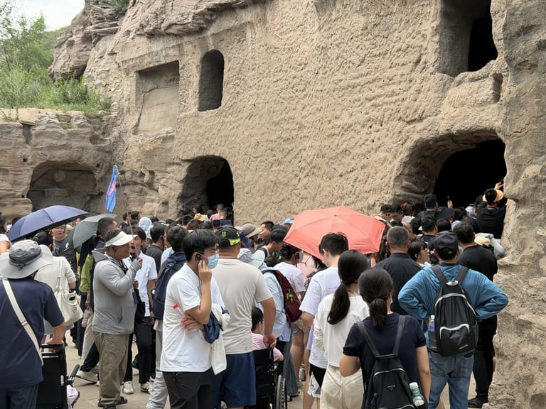 一二窟前挤满游客,一人举着红色阳伞,有人举着导游旗,岩壁上的洞口开在人群上方