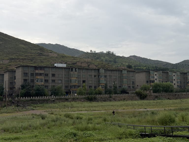 依山而建的长条形五层灰色砖混宿舍楼,外观陈旧带铁皮阳台,前景是杂草河滩草地,远处山顶隐约可见一座小亭子