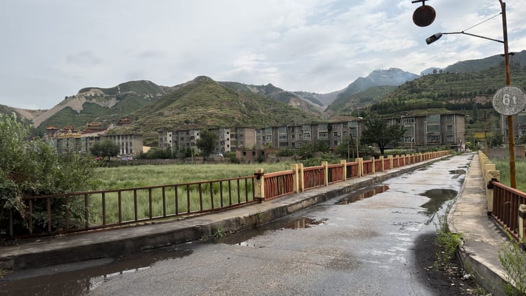 乡间水泥路横跨桥梁,桥面积水映出天空,桥两侧红褐色金属栏杆,远处是一排三四层灰色民居楼群,山脚下隐约可见一座带飞檐屋顶的庙宇