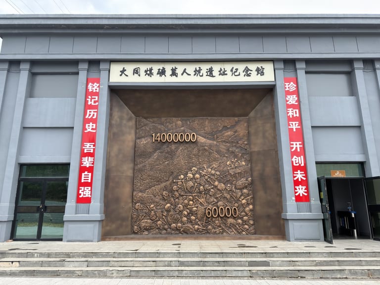 灰色建筑外墙上挂着白底黑字匾额上书「大同煤矿万人坑遗址纪念馆」,入口两侧各悬挂红色竖幅,左书「铭记历史 吾辈自强」,右书「珍爱和平 开创未来」