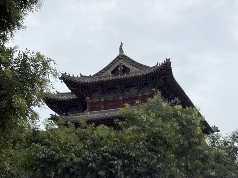 大同古城墙上一座传统中式楼阁的顶部,双层飞檐翘角,灰色筒瓦覆顶,檐下彩绘斗拱装饰,红色木窗紧闭,建筑被茂密绿叶簇拥