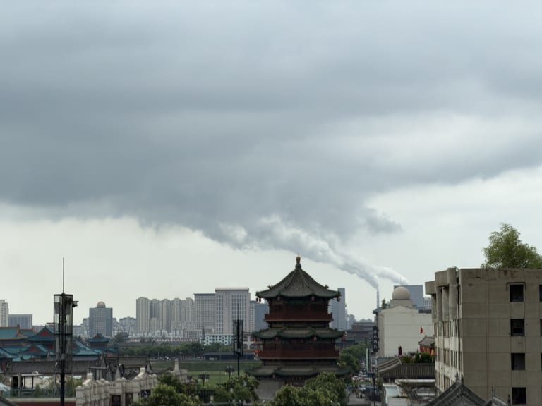 城墙外的中式多层建筑前矗立一座木质结构的高层阁楼,飞檐翘角青灰瓦顶金色宝顶;远方可见密集的高层住宅楼和冒着白烟的烟囱