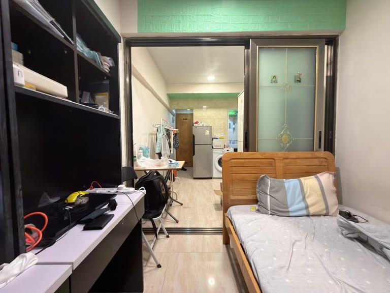 太子一间150呎唐楼单间内景——从床那一侧朝门口方向，左侧胡桃木书桌、右侧木床，正前方玻璃趟门外可见厨房的雪柜与洗衣机