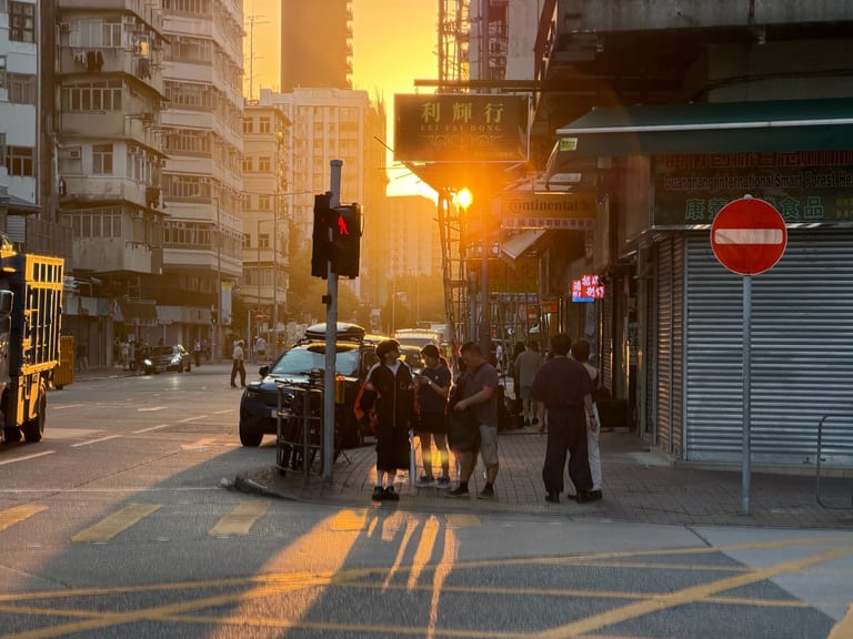 傍晚的香港街角,红绿灯前几个行人在等车,远处霓虹招牌"利輝行"在夕阳里发亮