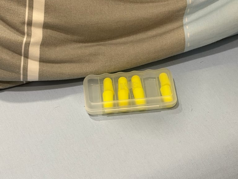 透明塑料药盒里四对黄色海绵耳塞，落在浅蓝灰床单和卡其色枕头上